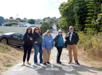 Five people standing on a narrow road with "STOP" painted on it, surrounded by trees and houses in the background.