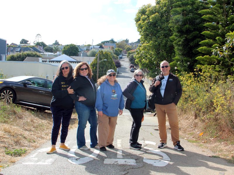 Five people standing on a narrow road with "STOP" painted on it, surrounded by trees and houses in the background.