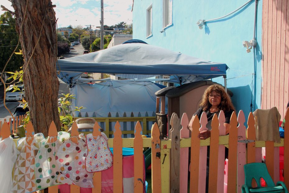 A woman stands behind a colorful wooden fence, with clothes hanging on it. A blue house and a canopy are in the background.