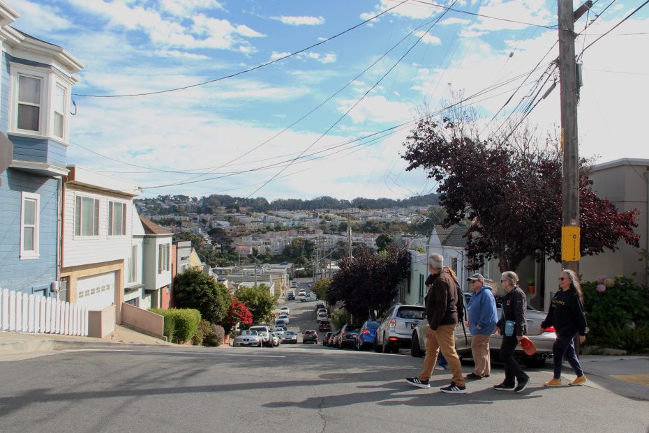 People walking down a sunny residential street, with houses on either side and a view of a distant neighborhood.