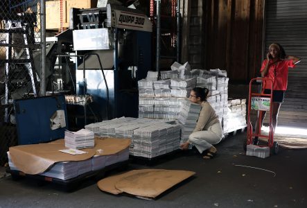 Two people in a warehouse with stacks of newspapers. One person is crouched by the stacks; the other stands next to a dolly and is on the phone. Machinery and a fence are in the background.
