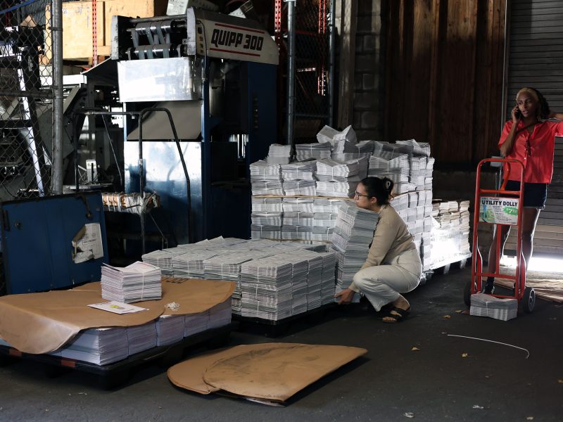 Two people in a warehouse with stacks of newspapers. One person is crouched by the stacks; the other stands next to a dolly and is on the phone. Machinery and a fence are in the background.