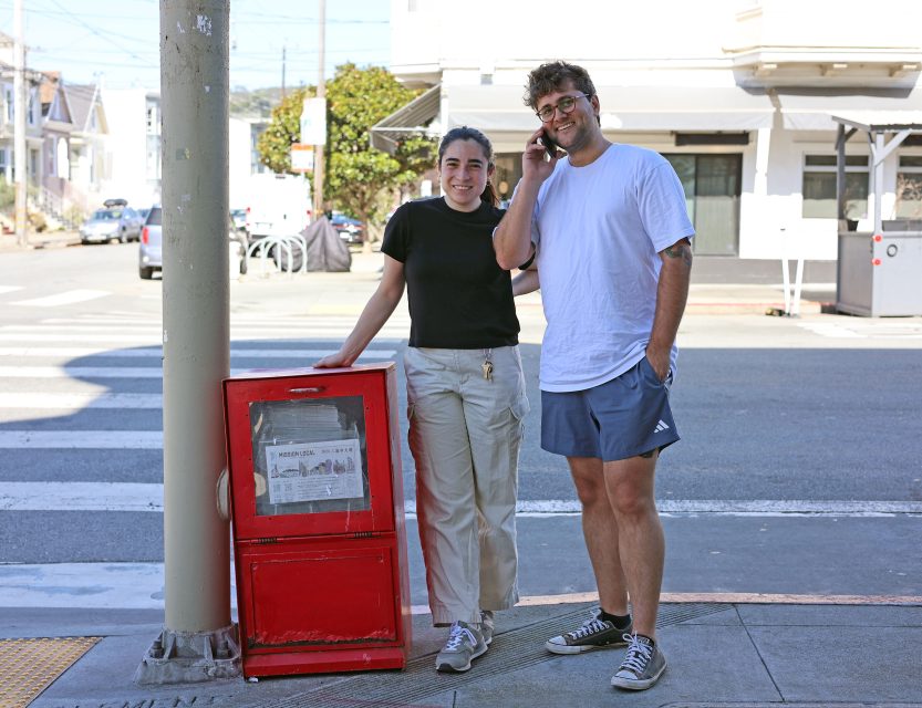 Two people standing on a street corner, leaning against a red newspaper box. One is on the phone, and both are casually dressed. A crosswalk and buildings are visible in the background.