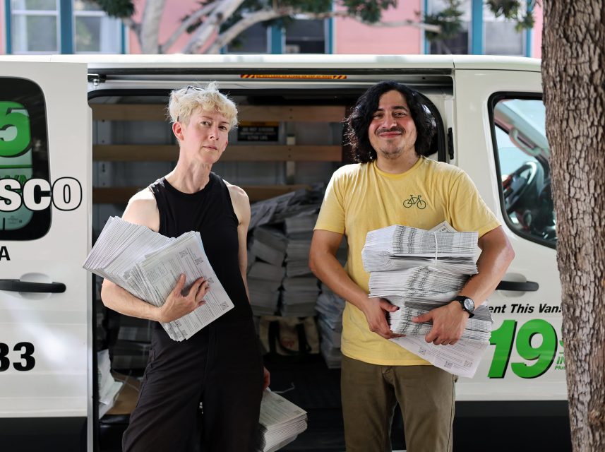 Two people standing in front of an open van holding stacks of newspapers.