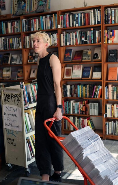 Person in black clothing pulls a cart stacked with newspapers in a bookstore with wooden shelves filled with books.