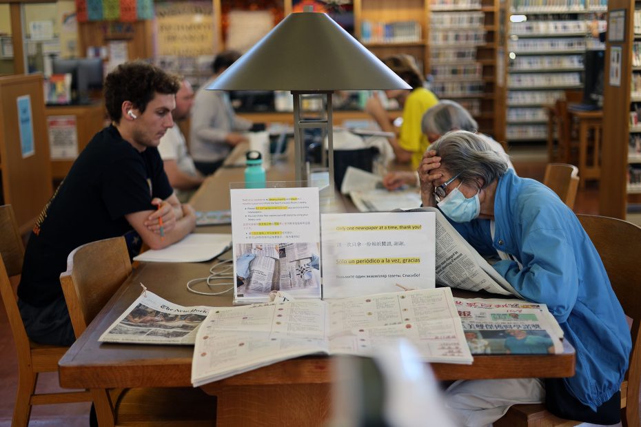 People reading and working at tables in a library; one person wears a mask and leans over reading a newspaper.