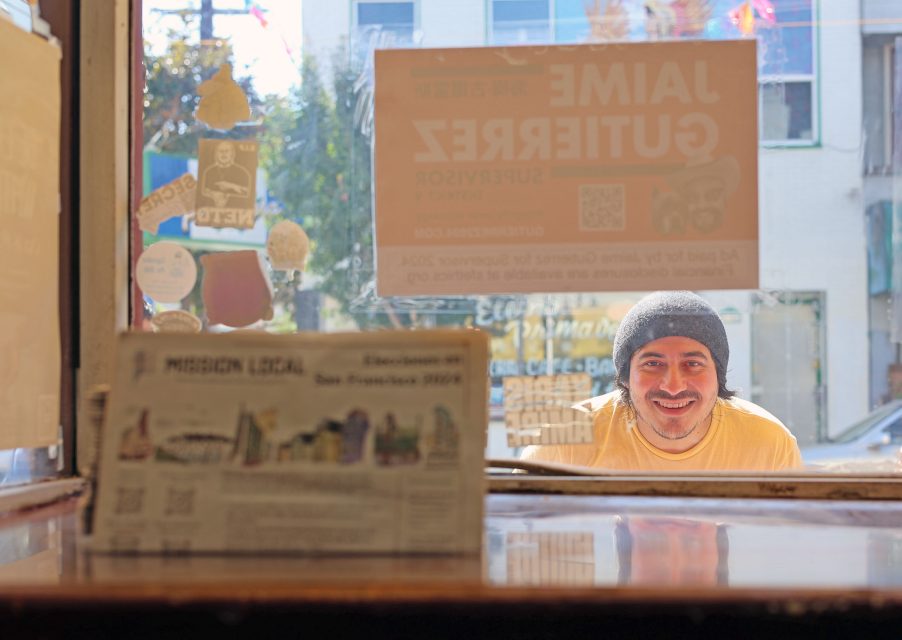 A smiling person wearing a beanie looks through a cafe window with newspapers and posters visible inside.
