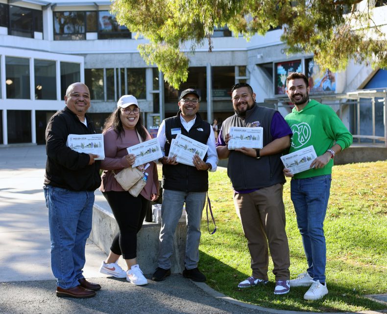 Five people standing outdoors, smiling, and holding maps or brochures in front of a modern building with large windows and a tree in the foreground.
