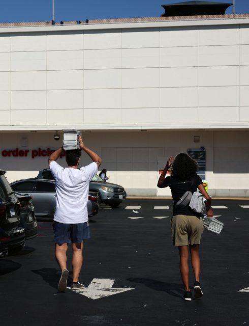 Two people carrying stacks of newspapers walk through a parking lot toward a large white building.