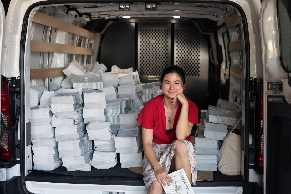 A person in a red shirt sits inside a van filled with stacks of newspapers, holding one in hand and smiling.