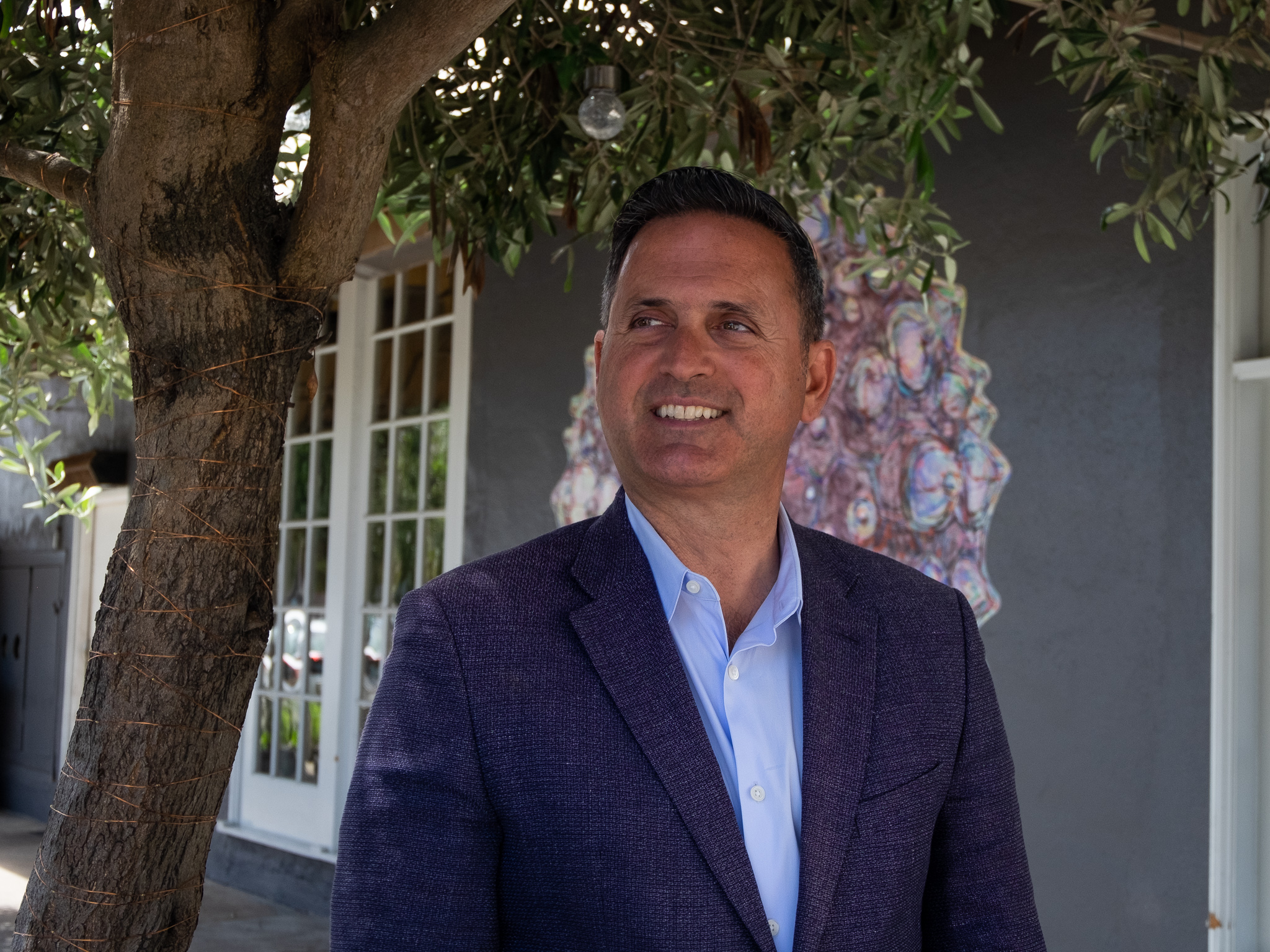 A person in a blazer stands under a tree, smiling, with a building in the background.