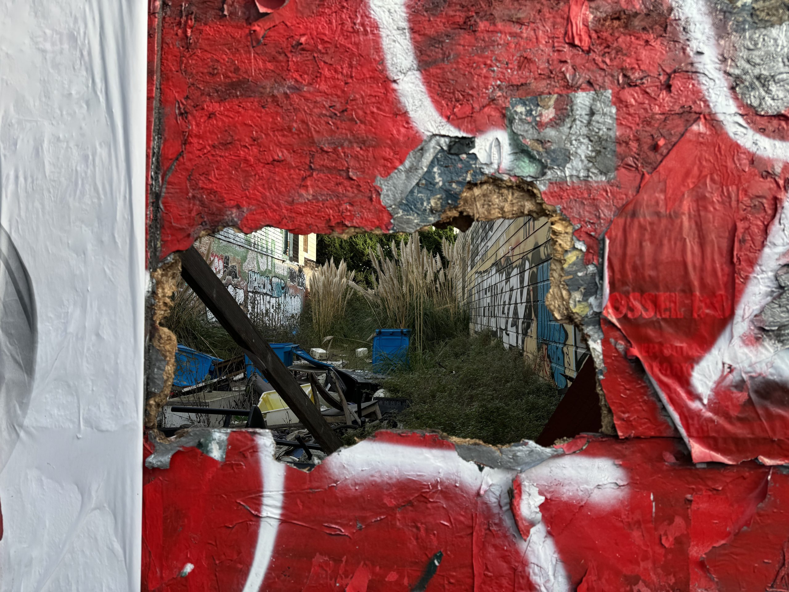 A hole in a red graffiti-covered wall reveals an overgrown outdoor area with blue containers and scattered debris.