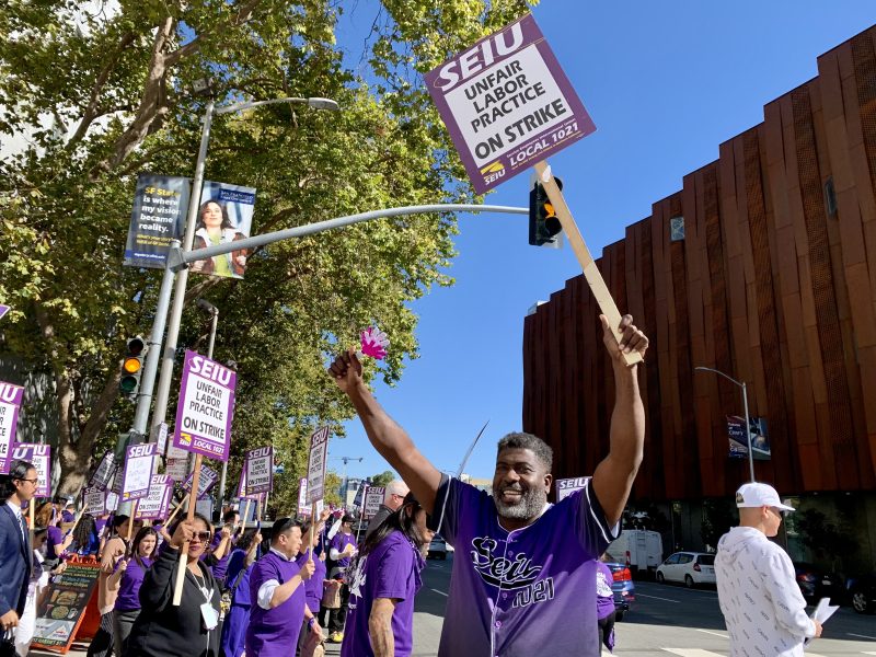 A group of people in purple shirts hold signs reading "SEIU Unfair Labor Practice On Strike" during a protest on a city street.