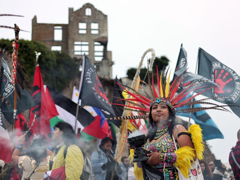 A person in traditional attire with a feathered headdress stands amid a crowd holding flags, with a ruined building in the background. Smoke is visible in the foreground.
