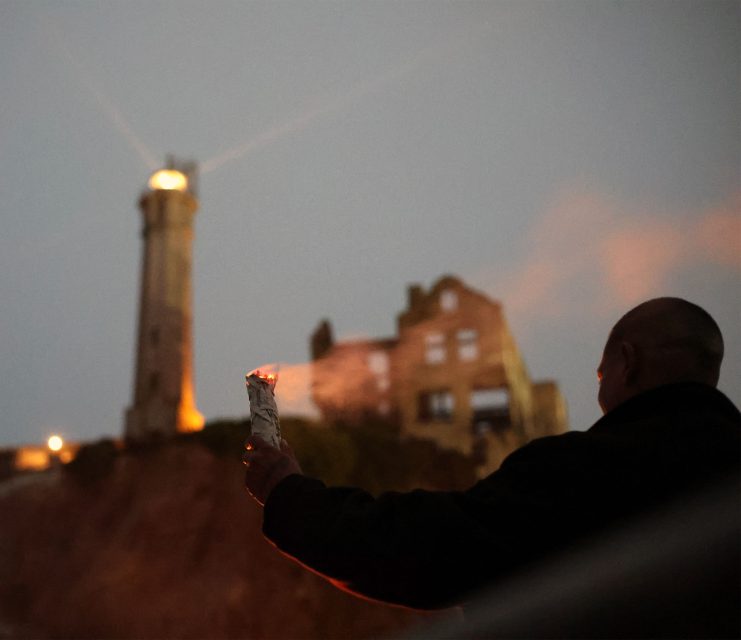 A person holding a burning paper stands in front of a lighthouse at dusk with an abandoned building nearby.