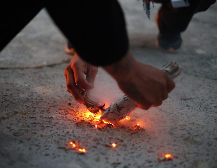 Hands holding a smoldering bundle of sage above a concrete surface, with small glowing embers creating smoke.