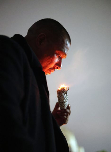 A man holding a burning sage bundle close to his face, with smoke rising, against a gray background.