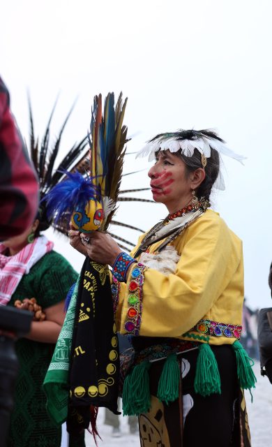 Person in traditional clothing holds colorful feathers, wearing a beaded yellow garment and headdress with feathers, face painted, standing outdoors.