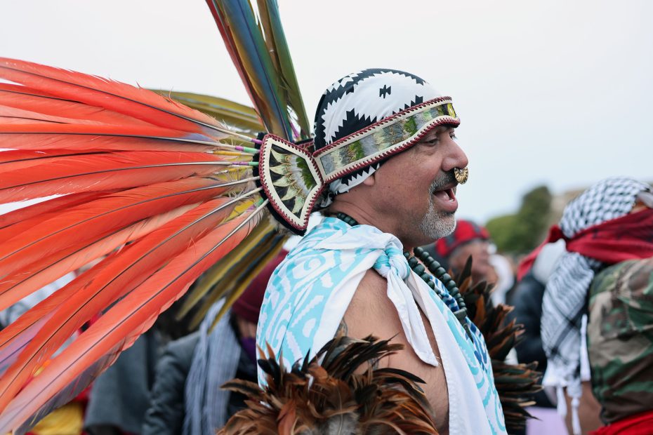 A person wearing a headdress with red and green feathers, patterned fabric, and a nose ring is participating in a cultural event.