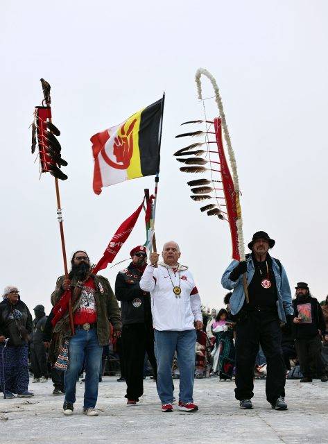 Three men hold flags with feathers, standing outdoors in winter clothing. A crowd is visible in the background.