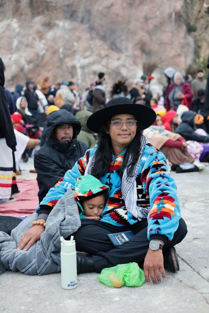 A person in a colorful jacket and hat sits on the ground, holding a child wrapped in blankets. There are people in the background wearing winter clothing.