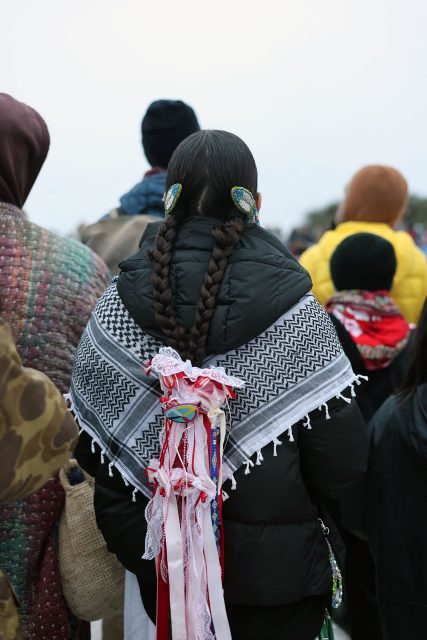 A person with braided hair, adorned with beaded hair accessories, wears a patterned shawl with red and white ribbons, standing among a group of people.