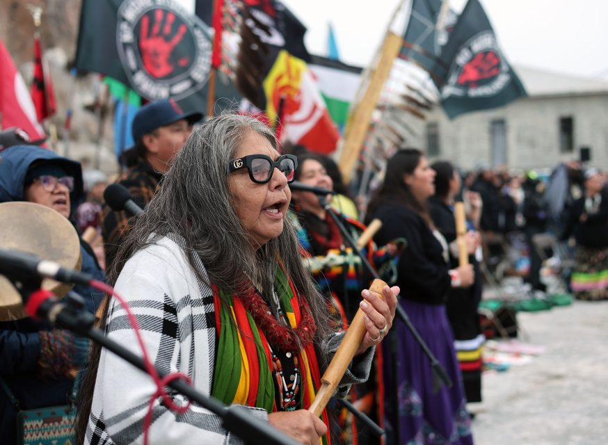 A vibrant group of people in colorful clothing stands outdoors near Alcatraz, holding musical instruments and flags. They seem to be part of a cultural event or gathering, celebrating their heritage with music and unity.