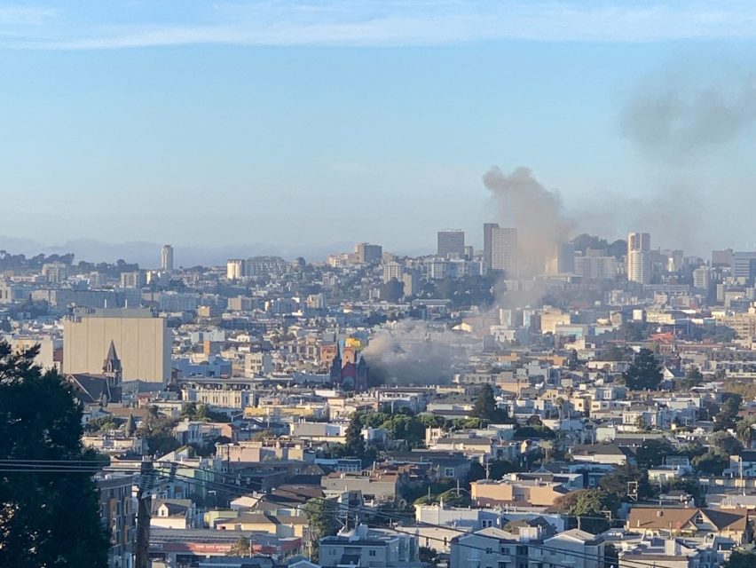 View of a cityscape with smoke rising from multiple locations, under a clear blue sky.