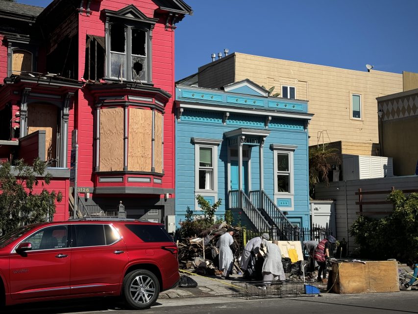 Two boarded-up houses, one red and one blue, with people sorting belongings on the sidewalk. A red SUV is parked in front.