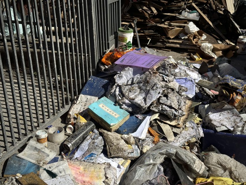 Pile of debris including torn papers, a blue book, and various discarded items next to a metal fence.