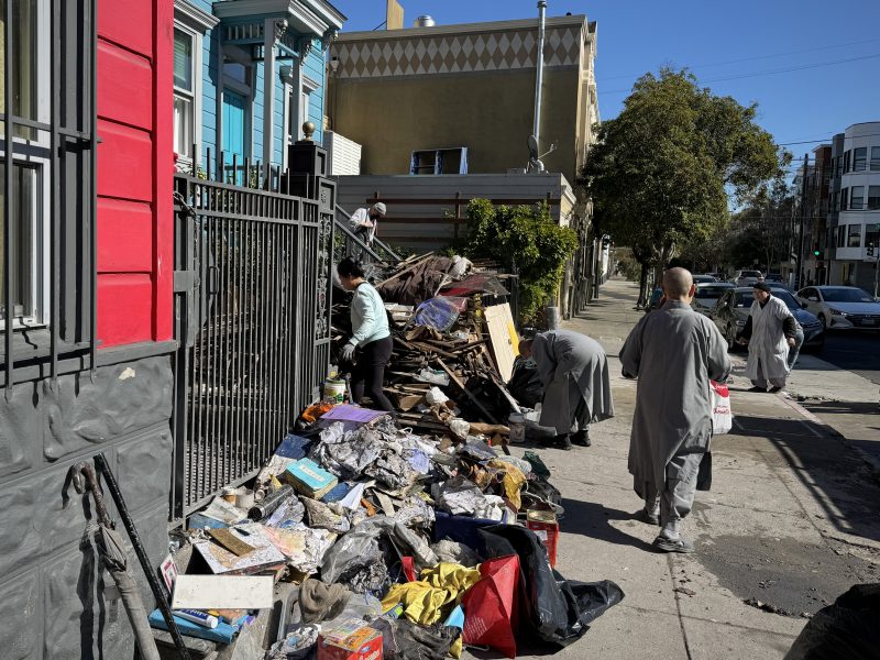 People in gray clothing clean up debris and garbage piled on a sidewalk beside a metal fence, with colorful buildings and trees in the background.