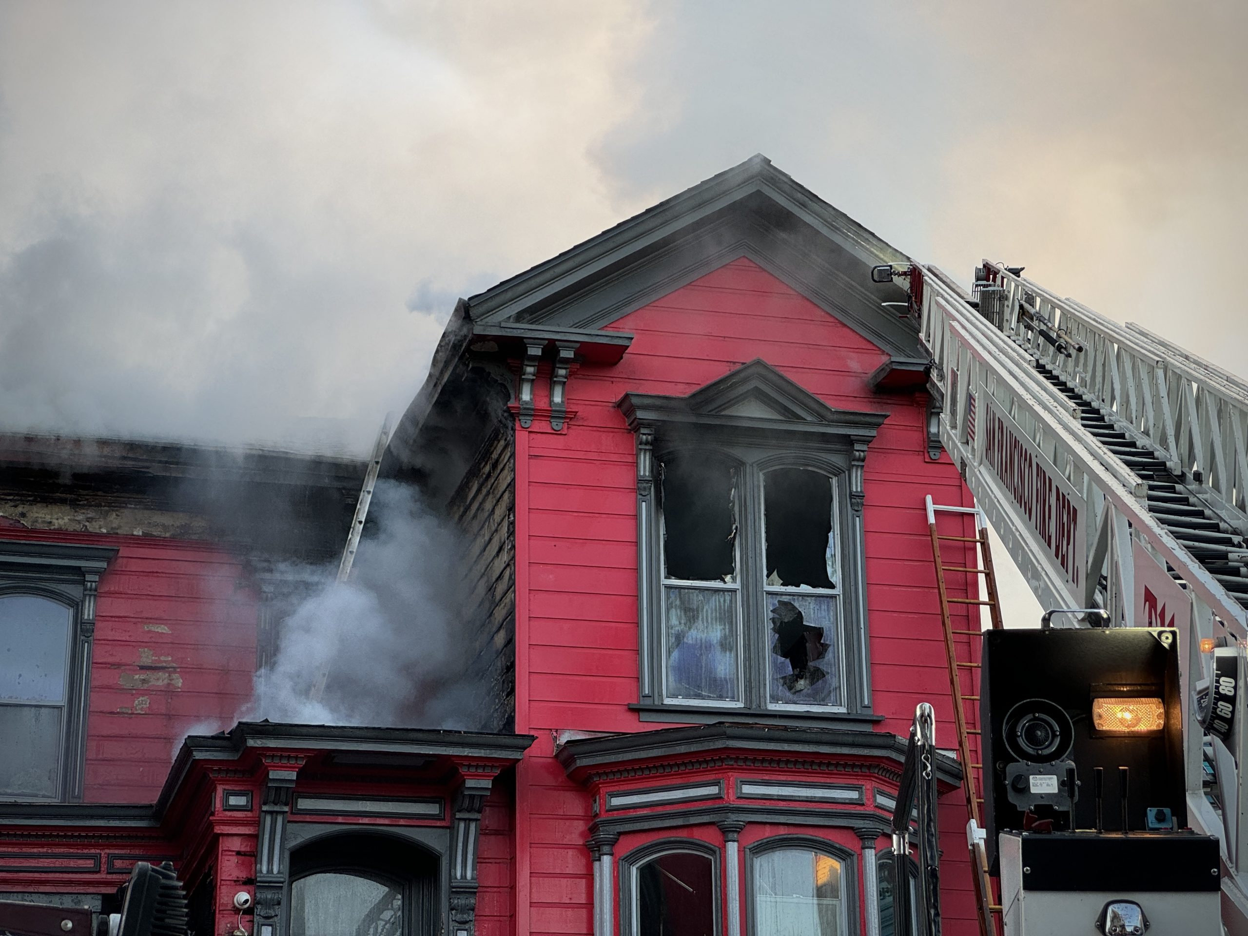 A red building on fire with smoke billowing out. Firefighters on a ladder work to control the blaze. Broken windows visible.