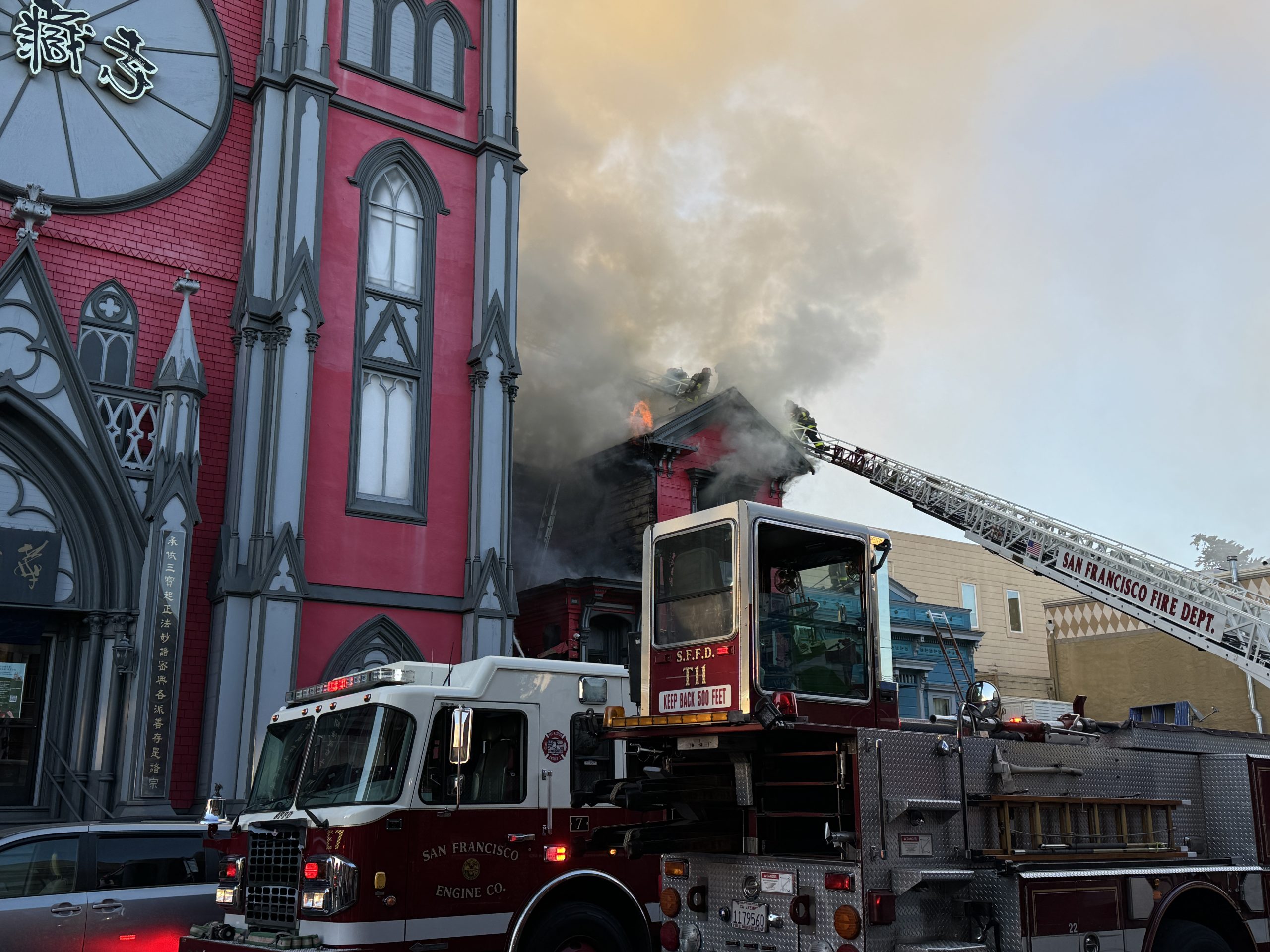 Firefighters respond to a building fire with a ladder truck outside a red and gray structure. Smoke and flames are visible from the roof.