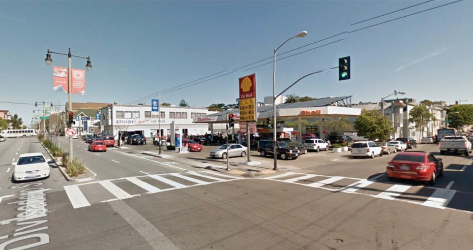 Intersection with a Shell gas station on one corner, cars stopped at a traffic light, and several buildings in the background under a clear blue sky.