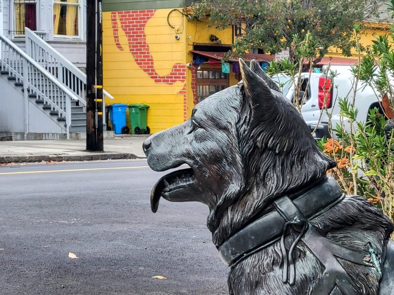 A dog statue with its tongue out sits near a street, with colorful buildings and plants in the background.
