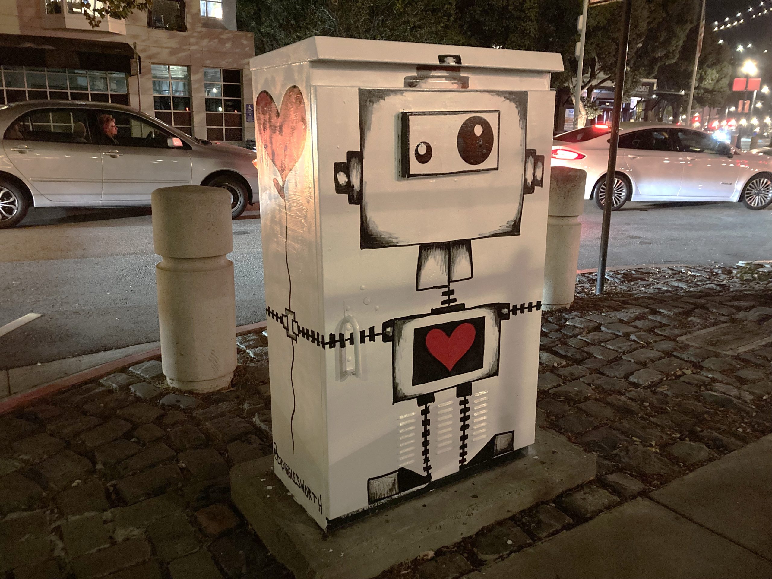 Utility box on a street corner painted with a robot design, featuring heart shapes on its body.