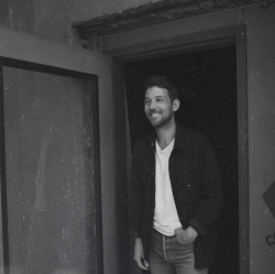 Black and white photo of a man standing in a doorway, smiling, with one hand in his pocket. He's wearing a dark jacket over a light shirt and jeans.