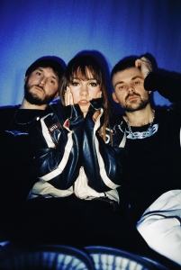 Three people posing against a blue-lit background, two men on either side of a woman. All are dressed casually and looking directly at the camera.