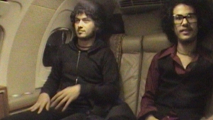 Two people sitting in an airplane cabin, wearing casual clothing and glasses.