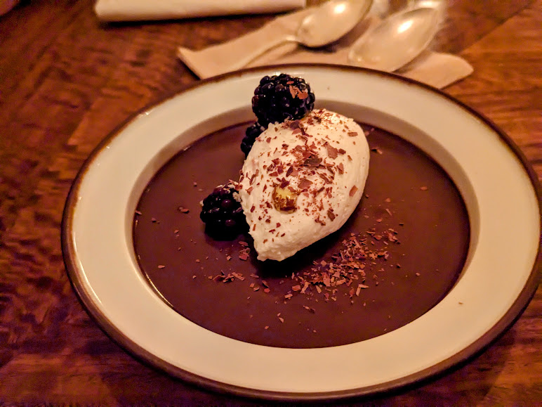 A dessert with a layer of chocolate mousse topped with whipped cream, blackberries, and chocolate shavings on a white plate.