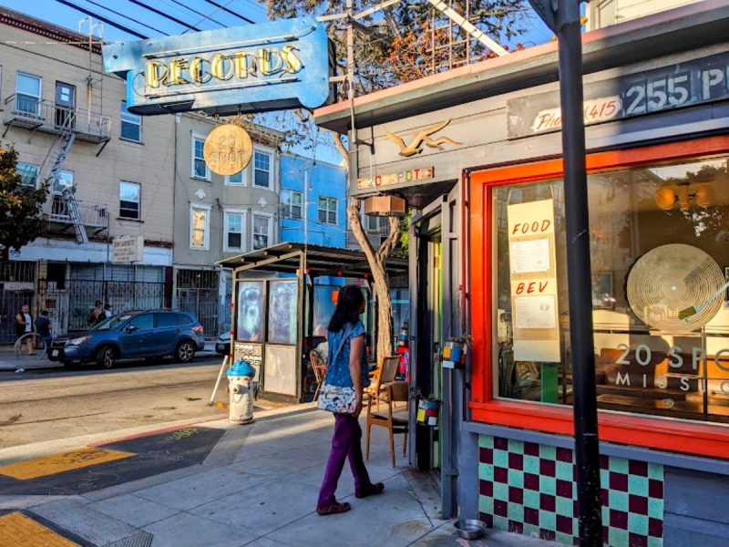 A person walks past a colorful record store and a food and beverage shop on a sunny city street corner.