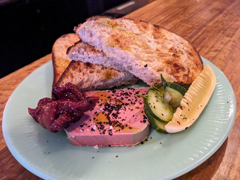 Plate with toasted bread, liver pâté topped with black pepper and oil, pickled cucumber, and a lemon wedge.