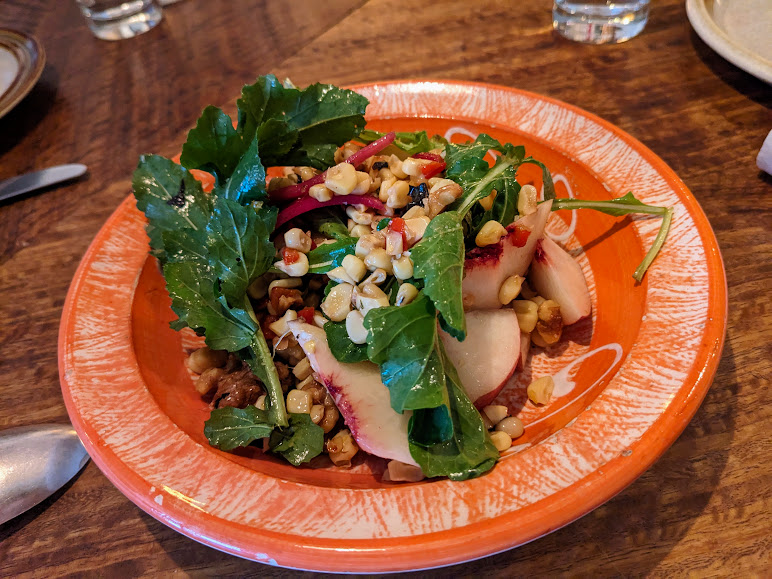 Salad with sliced peaches, arugula, walnuts, corn, and radishes on an orange plate.