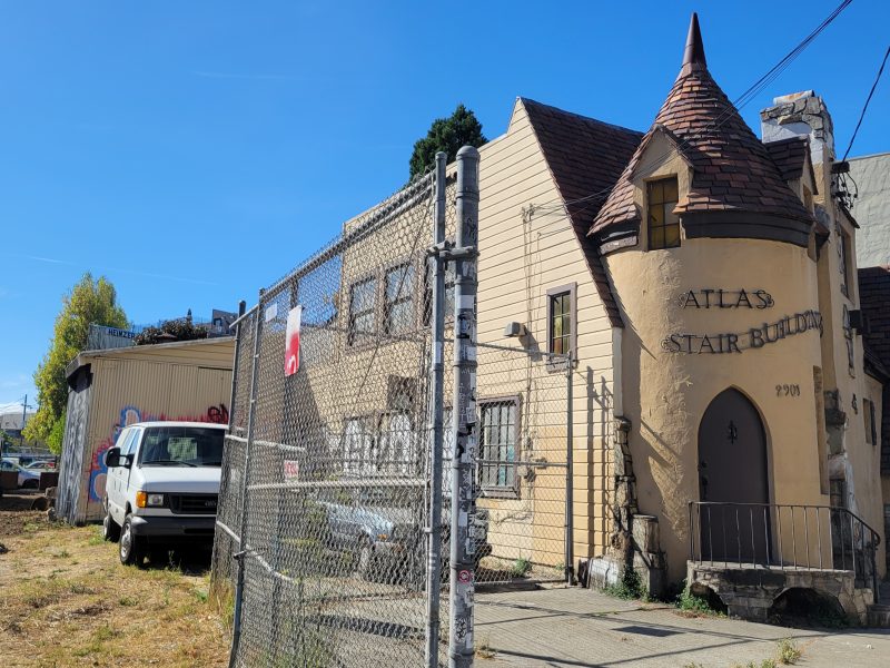 A fenced-off, castle-like building with a turret, labeled "ATLAS STAIR BUILDERS," alongside parked vehicles on a sunny day.