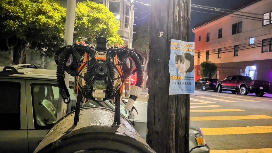 A homemade robot rests on a barrel by a street at night, next to a utility pole with a missing cat poster offering a reward.