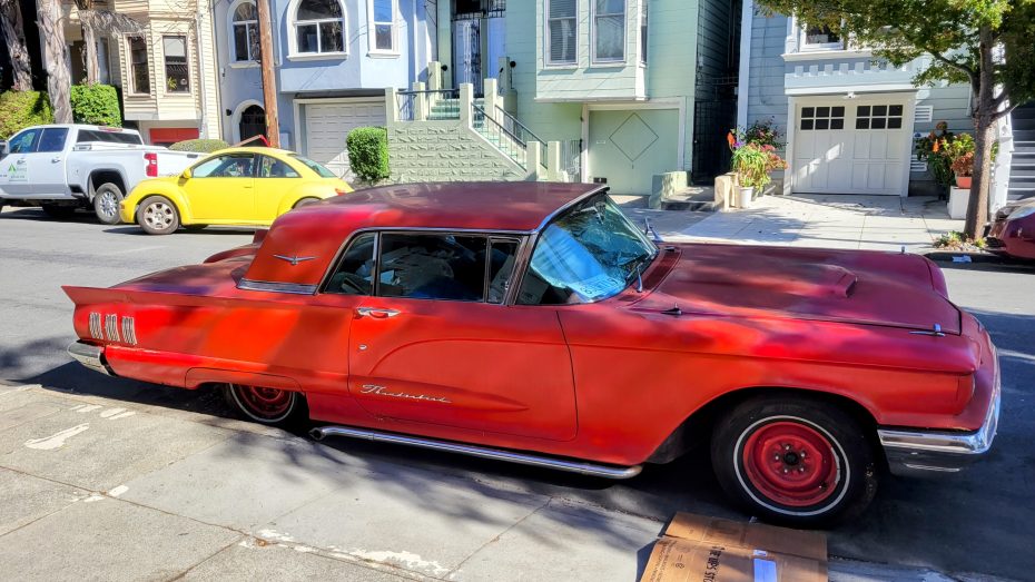 A red vintage car parked on a residential street, next to a yellow vehicle and in front of colorful houses with bay windows and a garage.