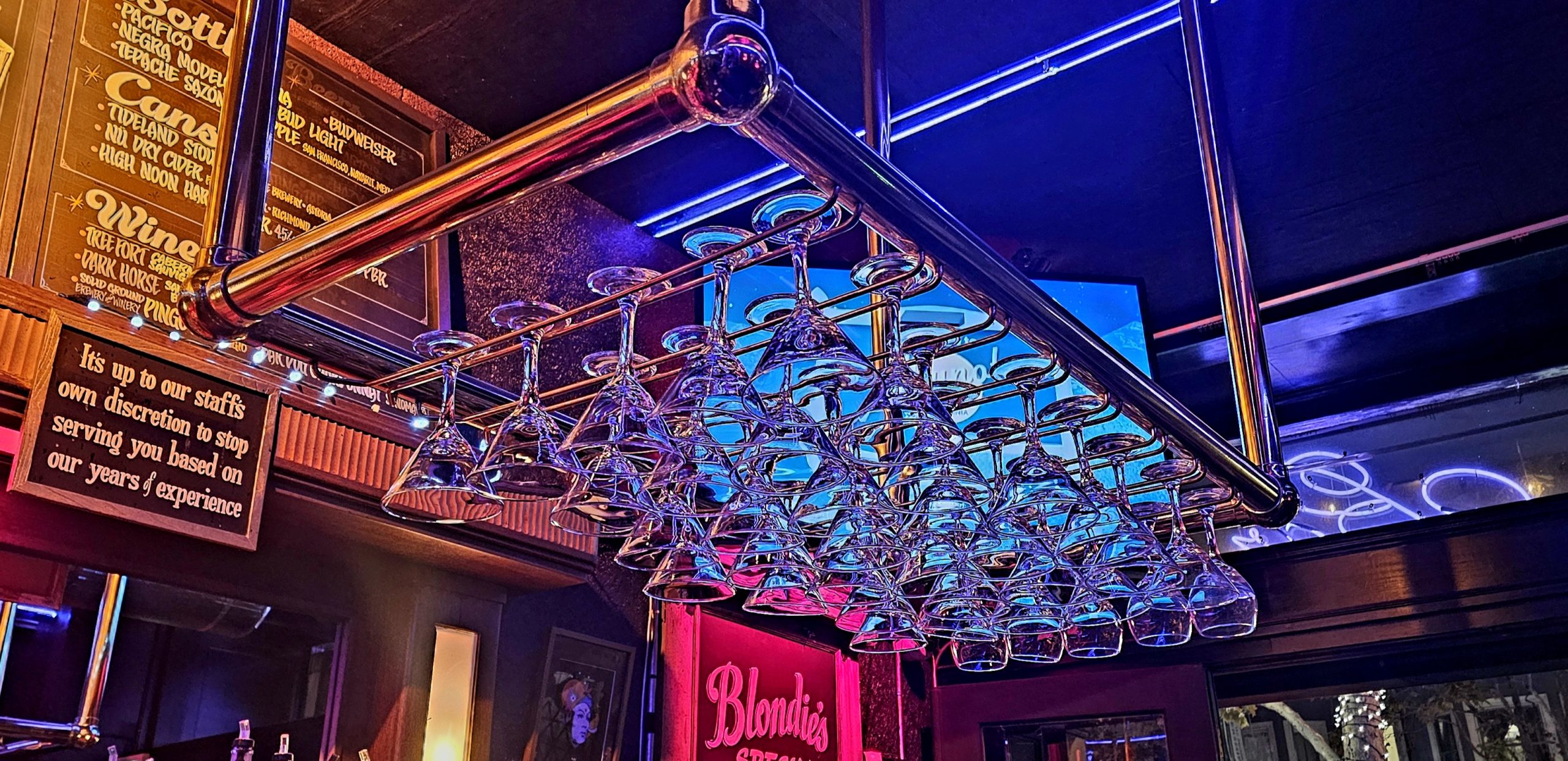 Rows of inverted glasses hanging from a rack in a dimly lit bar with neon lights and colorful signs in the background.