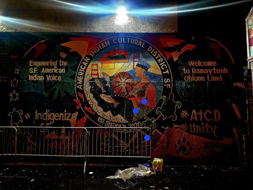 Mural of the American Indian Cultural District in San Francisco with symbolic elements, text about empowerment and unity, and cityscape imagery.