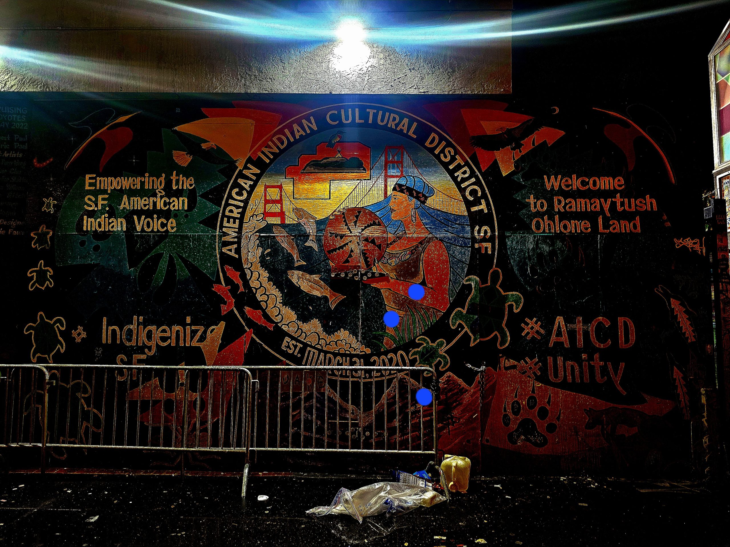 Mural of the American Indian Cultural District in San Francisco with symbolic elements, text about empowerment and unity, and cityscape imagery.