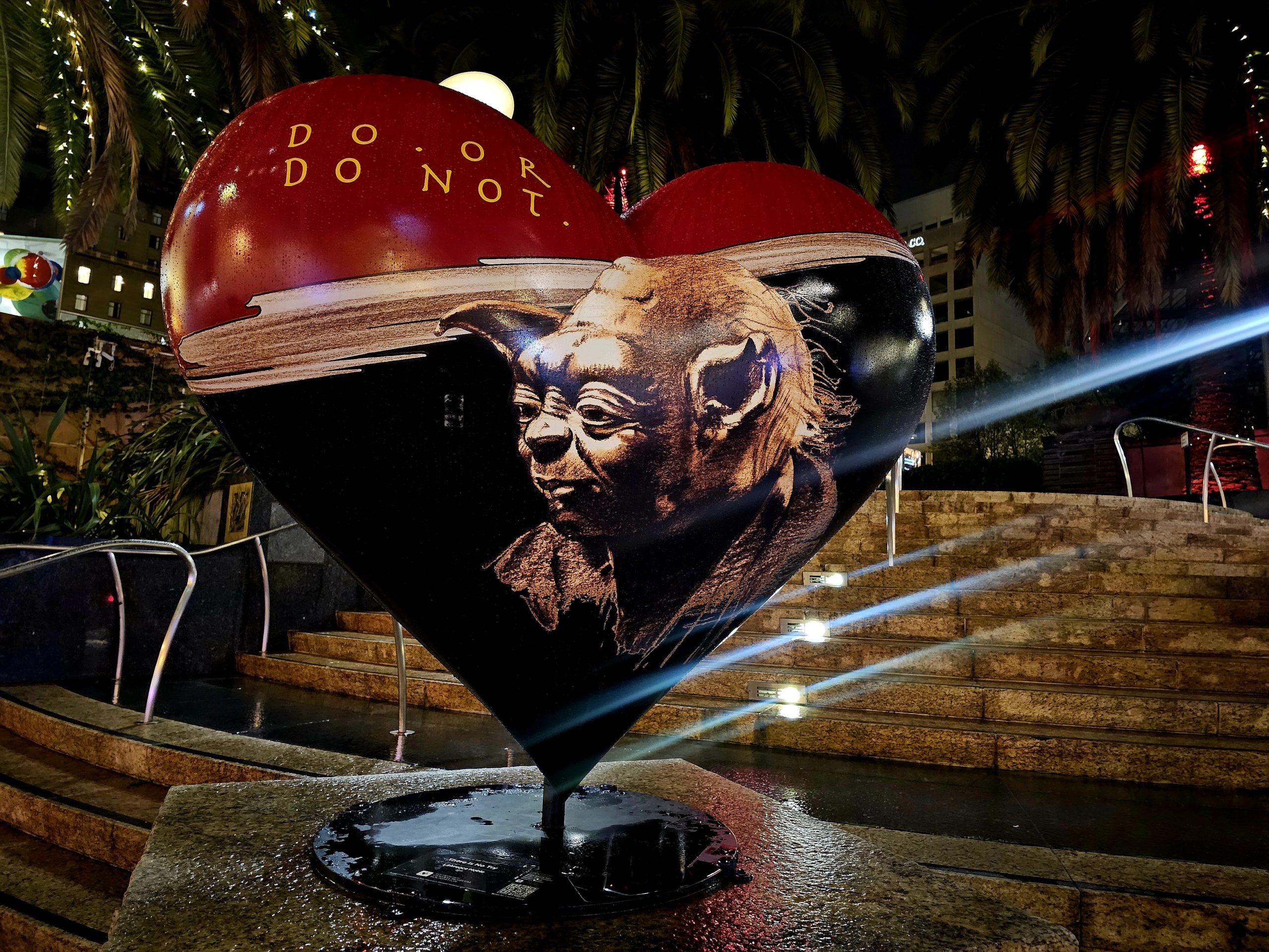 Heart sculpture in a city square, featuring Yoda from Star Wars with the phrase "Do or do not." Surrounded by steps and palm trees, illuminated at night.